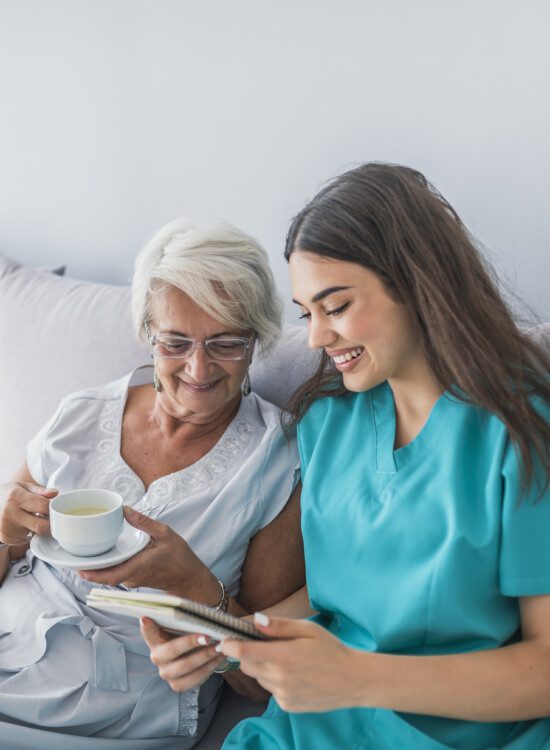 Happy patient is holding caregiver for a hand while spending time together. Elderly woman in nursing home and nurse. Aged elegant woman and tea time at nursing home