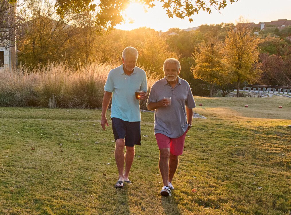 two casually dressed senior men carrying cocktails walk in the grass at sunset at Querencia Senior Living Community