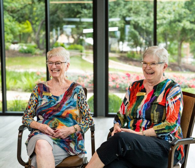 two smiling senior women, the Bain sisters, in colorful tops sit for an interview at Querencia Senior Living Community