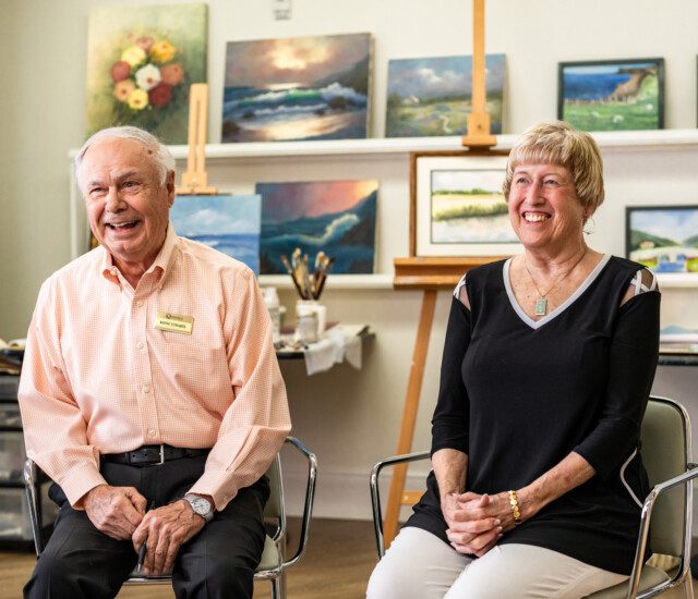 smiling senior couple sits for an interview, backdropped by a lineup of colorful paintings in the art studio at Querencia