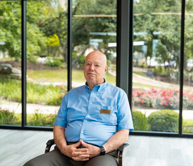 smiling senior man, Jev Sikes, in blue button-up shirt sits for an interview with hands folded in his lap, backdropped by large windows