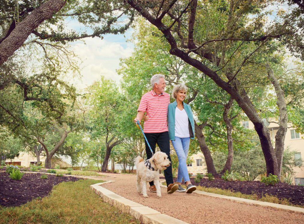 smiling senior couple with arms around one another walk their small white dog along a tree-lined path at Querencia Senior Living Community