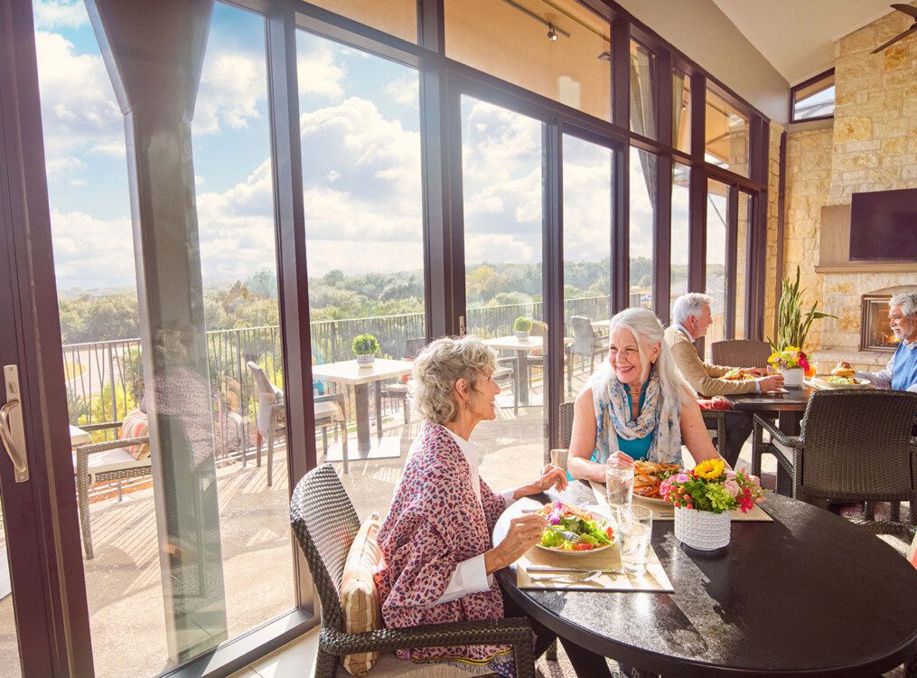 two tables of smiling, well-dressed seniors enjoy a meal in the dining room at Querencia, featuring a fire place and panoramic views