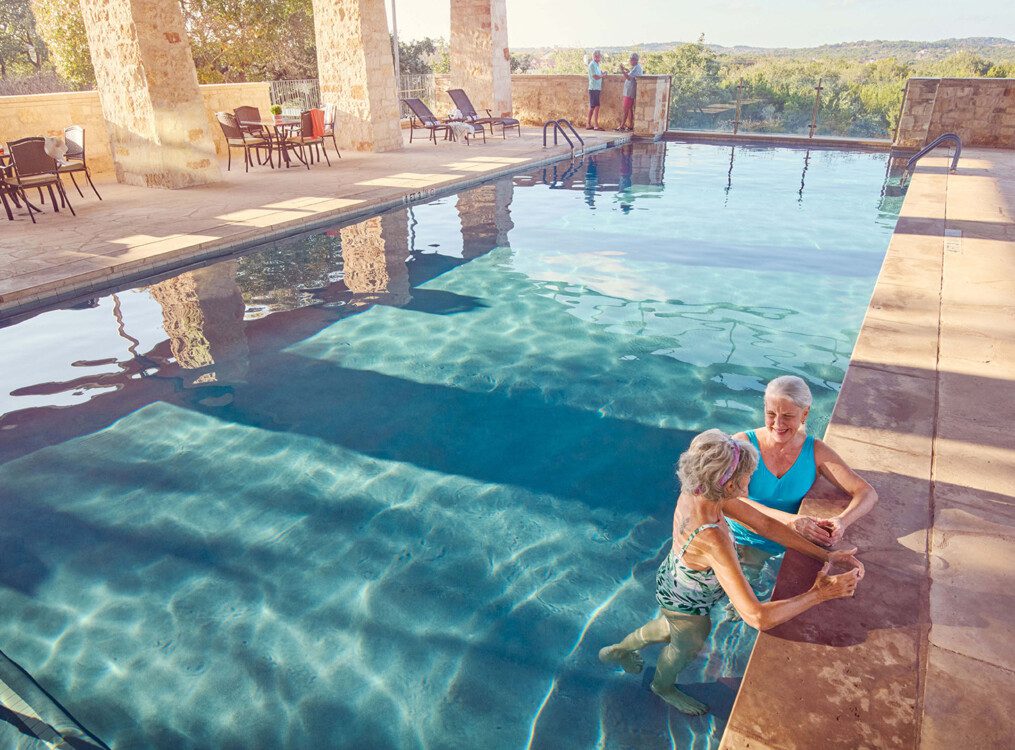 two senior women stand in the gorgeous outdoor swimming pool at Querencia Senior Living Community, leaning against its edge, enjoying drinks