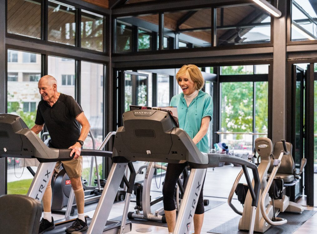 senior woman in athletic clothing smiles while walking on a high-tech treadmill at the fitness center at Querencia Senior Living Community