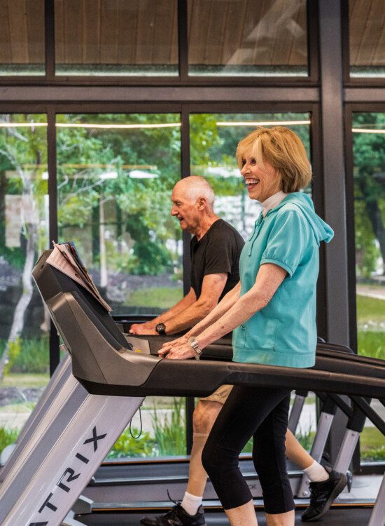 senior woman in athletic clothing smiles while walking on a high-tech treadmill at the fitness center at Querencia Senior Living Community