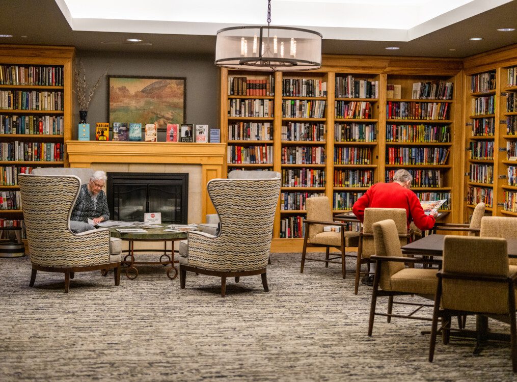 Timeless-feeling, traditional library at Querencia with fireplace, built-in bookshelves, seating, and two seniors reading