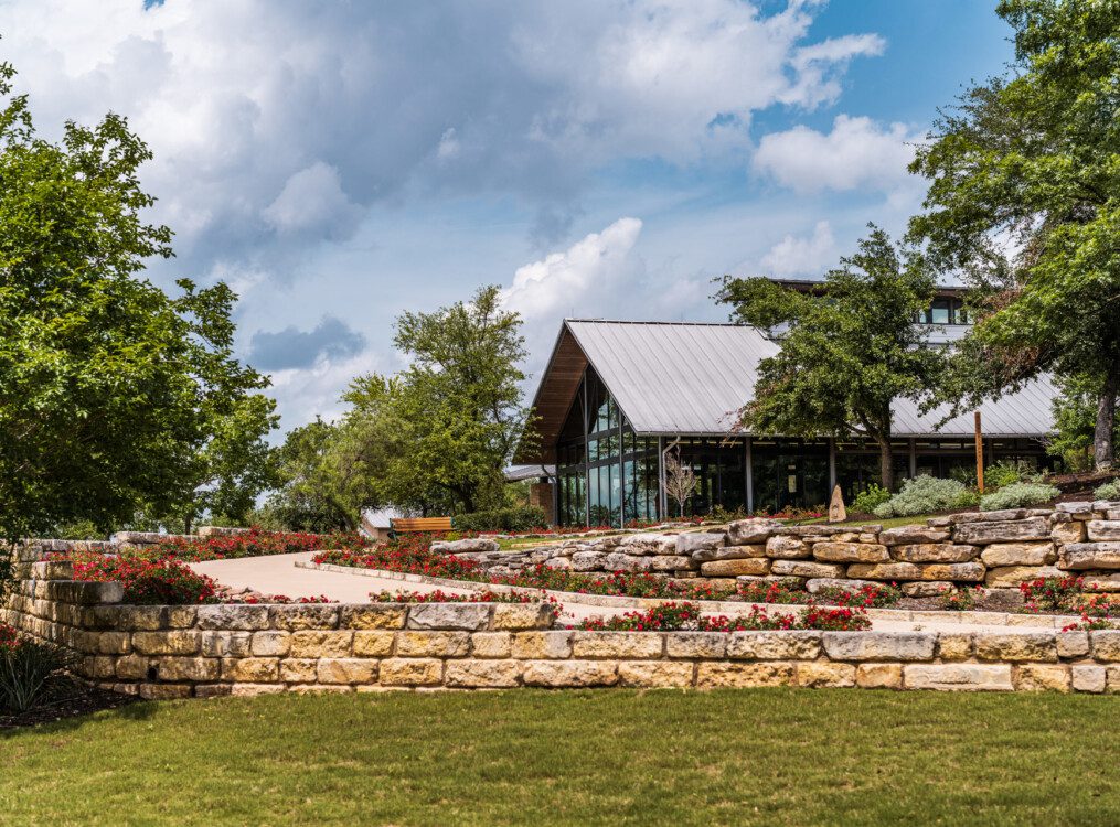 lush landscaping and stone along a driveway leading up to the glass fitness building at Querencia Senior Living Community in Austin, TX