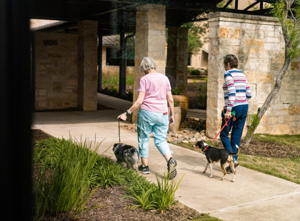 two senior women walk their dogs along a sidewalk at Querencia Senior Living Community in Austin, TX
