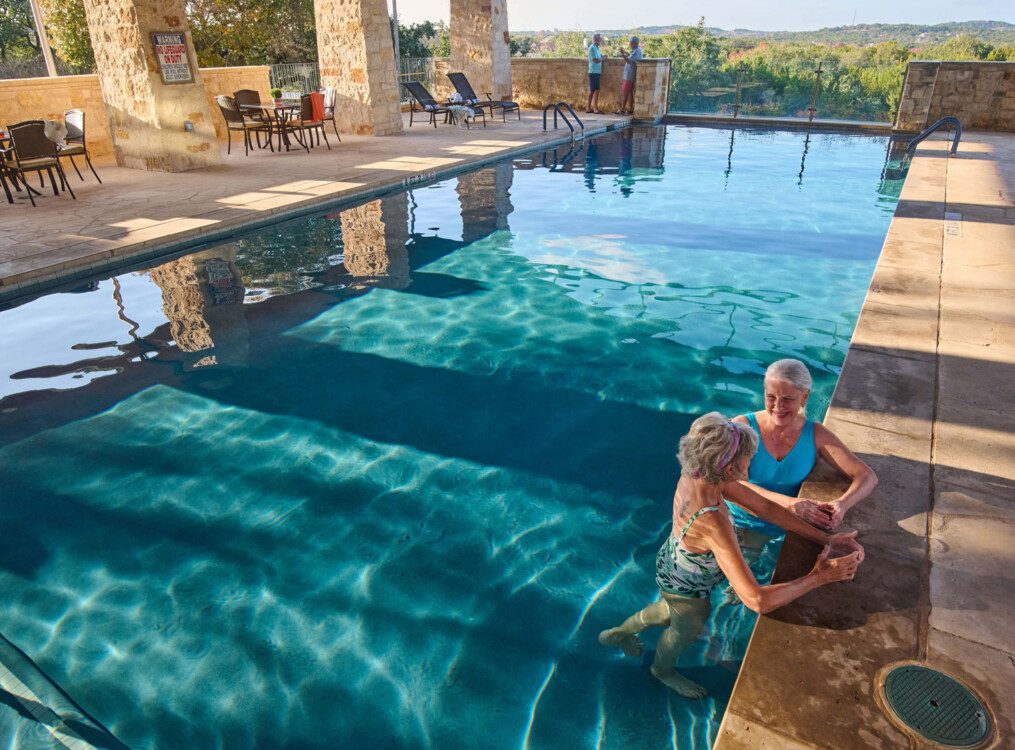 two senior women stand in the gorgeous outdoor swimming pool at Querencia Senior Living Community, leaning against its edge, enjoying drinks