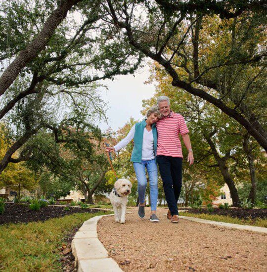 smiling senior couple with arms around one another walk their small white dog along a tree-lined path at Querencia Senior Living Community