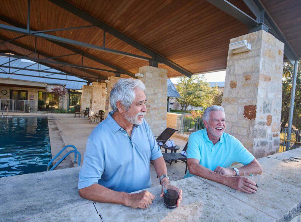 two casually dressed, smiling senior men enjoy drinks and great views along the stone edge of the outdoor lounge and pool area at Querencia