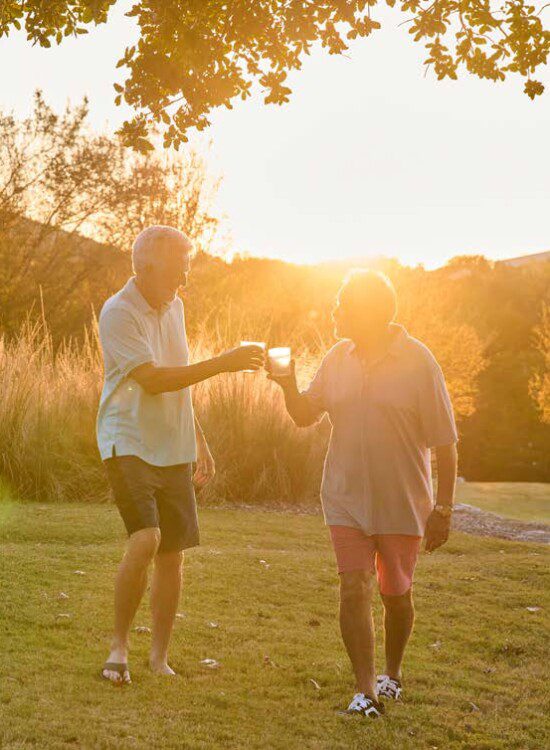 two senior men in casual clothing clink drinks together while walking outdoors, backdropped by the setting sun