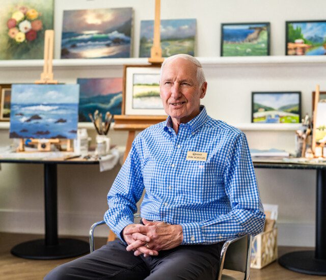 Well-dressed senior man, Thomas Schultz, sits in front of a collection of paintings in a studio during an interview