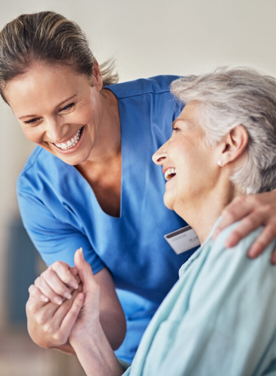 seated senior woman smiles up at her female caregiver, who is smiling with one arm around the woman's shoulder