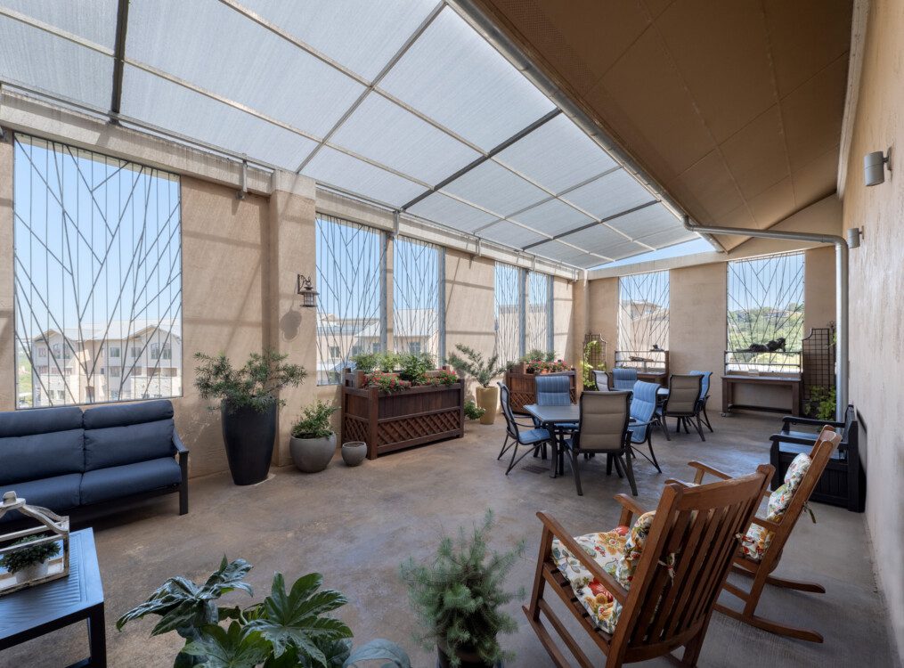spacious sunroom with multiple tables, rocking chairs, plants, and large windows at Querencia Senior Living Community's Assisted Living